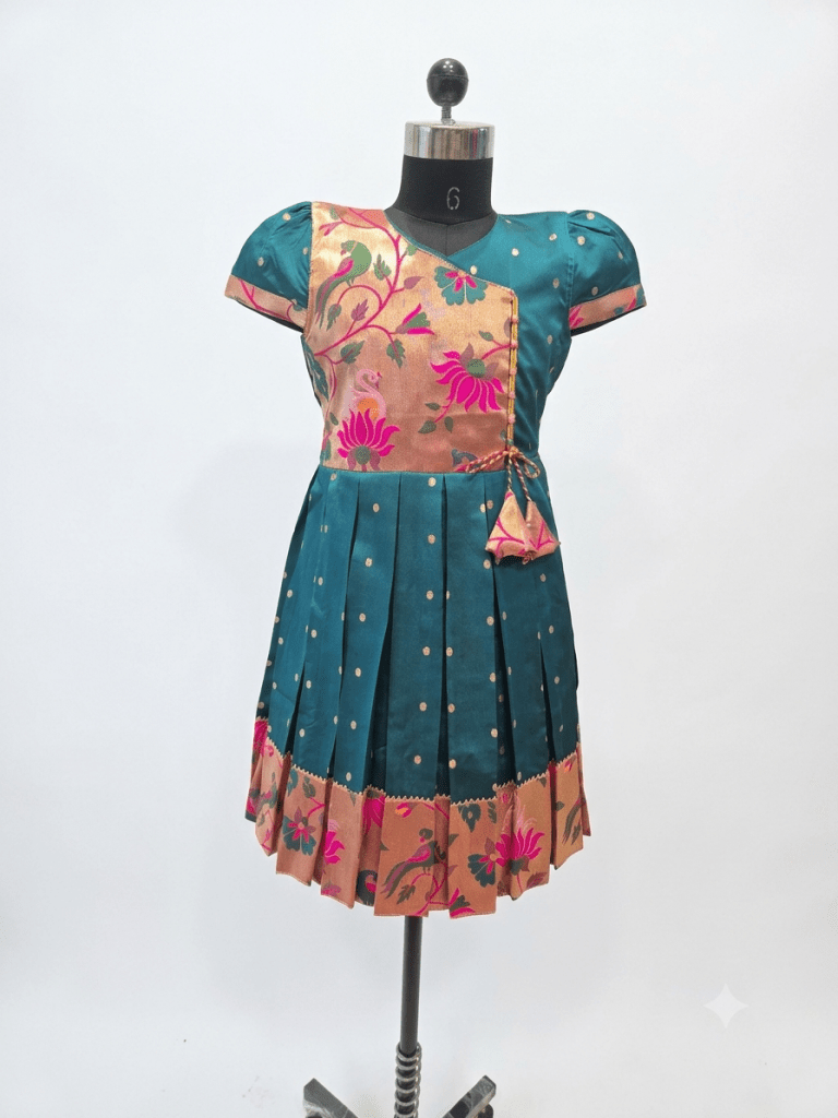 Young girl smiling in a traditional teal and gold silk pleated frock with floral embroidery, festive attire for kids.
