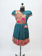 Young girl smiling in a traditional teal and gold silk pleated frock with floral embroidery, festive attire for kids.