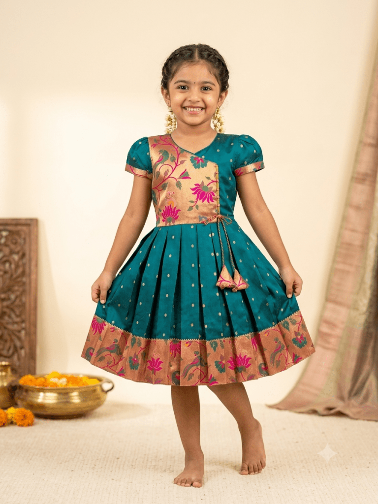Young girl smiling in a traditional teal and gold silk pleated frock with floral embroidery, festive attire for kids.