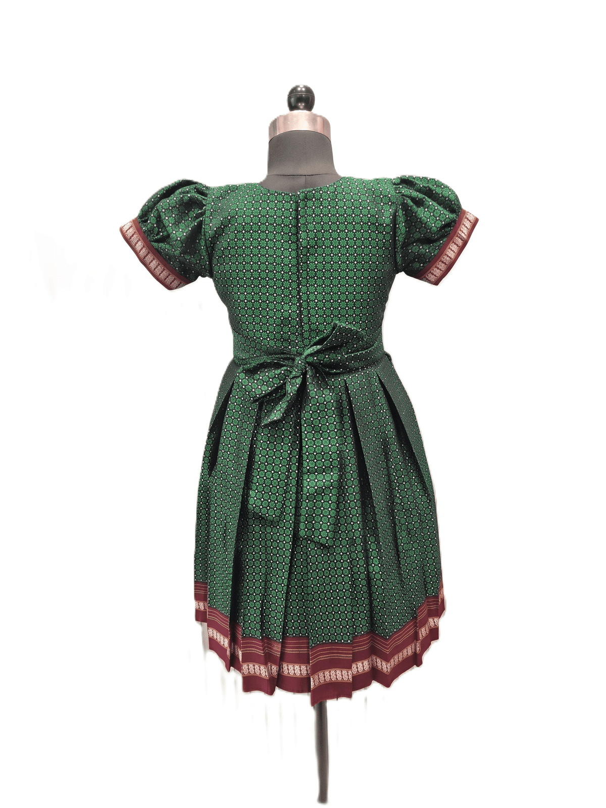 A green patterned dress displayed on a dress form mannequin against a white background. The dress features short puff sleeves with red and white patterned cuffs, a matching waist tie, and a pleated skirt with a red and white patterned hem. - House of Kritee
