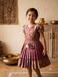 A young girl smiling and wearing a traditional Indian salwar kameez dress in a warm-toned indoor setting. The dress is a dusty rose/mauve color with gold zari embroidery and patterned details, featuring puff sleeves, a buttoned yoke, and a pleated skirt. - House of Kritee