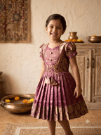 A young girl smiling and wearing a traditional Indian salwar kameez dress in a warm-toned indoor setting. The dress is a dusty rose/mauve color with gold zari embroidery and patterned details, featuring puff sleeves, a buttoned yoke, and a pleated skirt. - House of Kritee