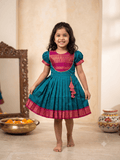 Happy young girl wearing a premium teal blue and magenta silk knee-length frock inside a beautifully decorated warm cream home - House of Kritee