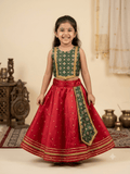 Young smiling Indian girl model wearing a traditional red and green silk parkar-polka lehenga choli set against a warm ivory ethnic background.