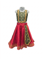 Young smiling Indian girl model wearing a traditional red and green silk parkar-polka lehenga choli set against a warm ivory ethnic background.