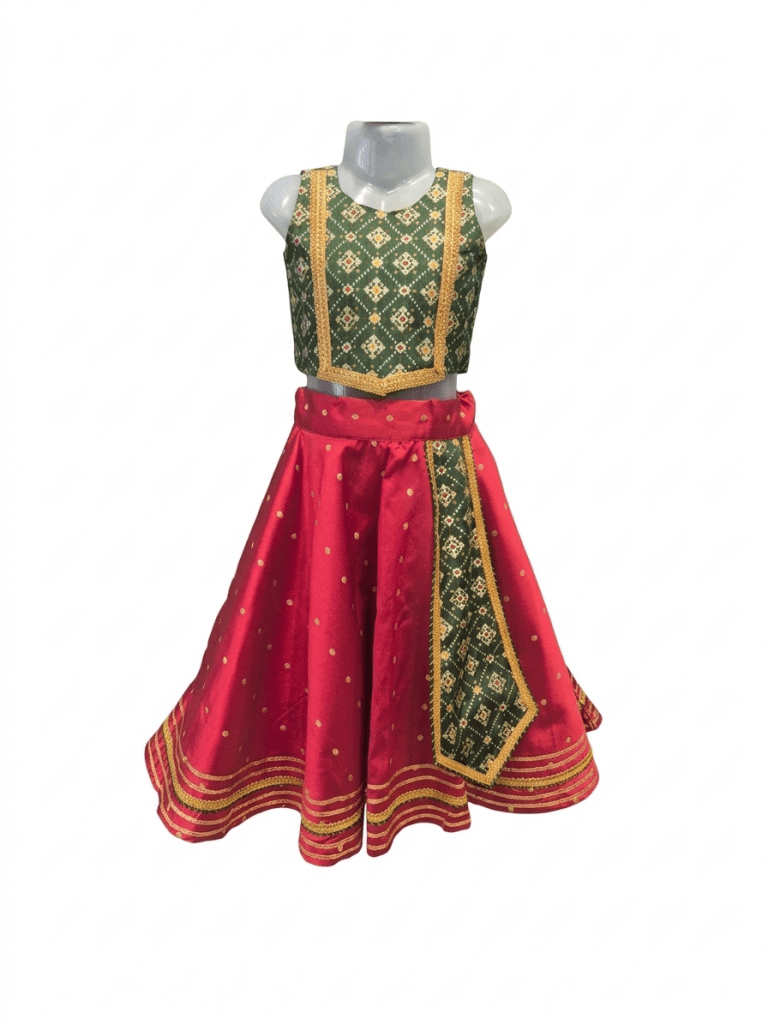 Young smiling Indian girl model wearing a traditional red and green silk parkar-polka lehenga choli set against a warm ivory ethnic background.