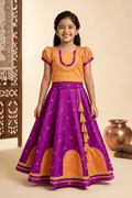 A young girl smiles while wearing a traditional orange and purple silk blend lehenga choli, featuring intricate pearl and gold embroidery and a decorative tassel